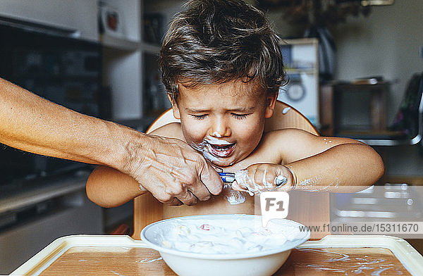 Little boy eating yogurt at home  hand of woman on the spoon