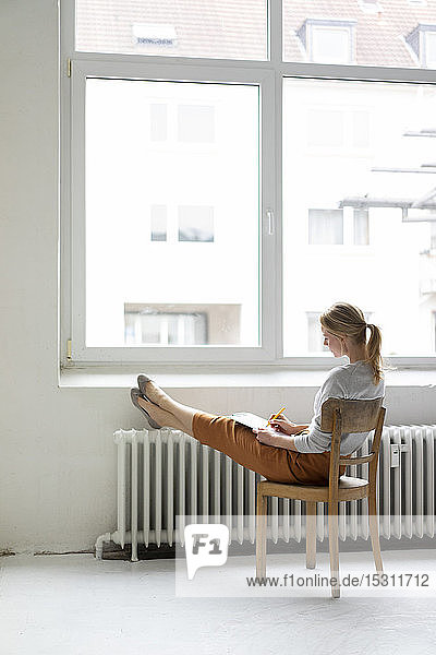 Junge Frau sitzt auf Stuhl am Fenster im Büro und macht Notizen