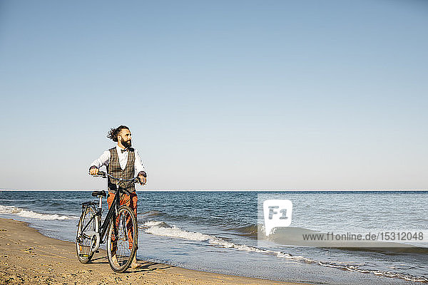 Gut gekleideter Mann geht mit seinem Fahrrad am Strand spazieren