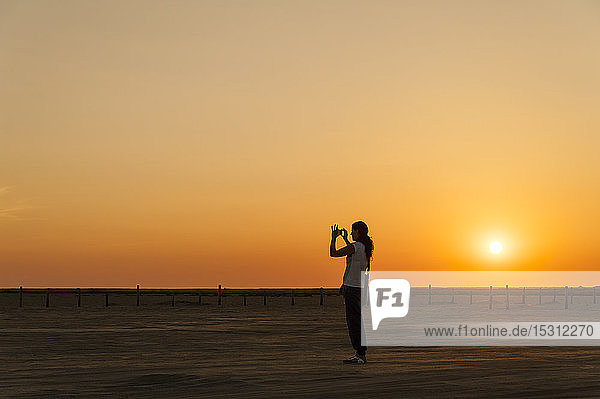 Deutschland Schleswig-Holstein Sankt Peter-Ording Frau beim Fotografieren am Strand bei Sonnenuntergang