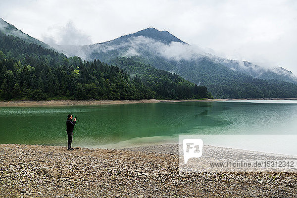 Man standing at lakeshore taking a photo with his smartphone  Wiestal reservoir  Salzburg State  Austria