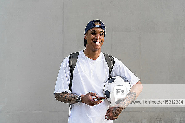 Portrait of tattooed young man with football and smartphone in front of concrete wall