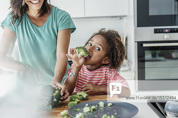 Girl cooking with mother in kitchen tasting broccoli