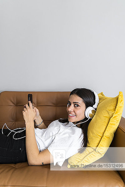 Junge Frau liegt zu Hause auf der Couch mit Smartphone und Kopfhörern