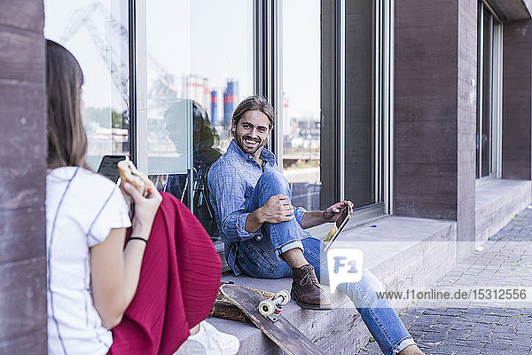 Lächelnder junger Mann sitzt auf der Fensterbank eines Gebäudes und schaut eine Frau an
