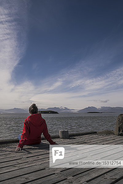 Junge Frau sitzt auf einem Steg und schaut auf das Meer  Südost-Island