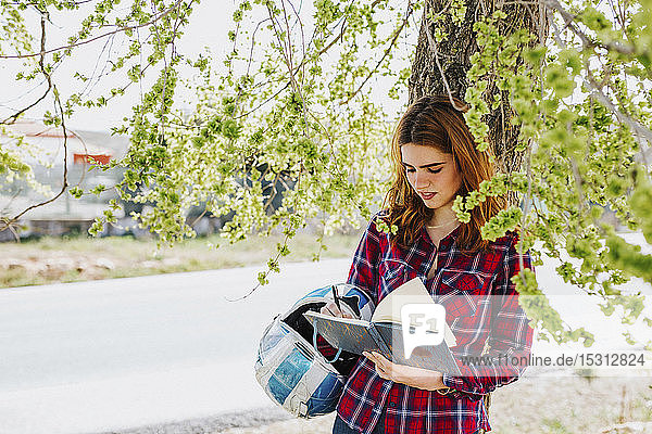 Redheaded woman with motorcycle helmet leaning against tree taking notes