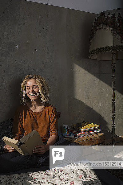 Porträt einer lachenden jungen Frau  die auf dem Bett sitzt und ein Buch liest