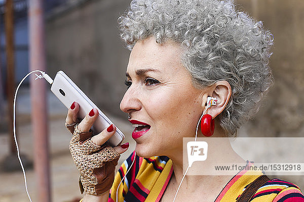 Portrait of pierced mature woman with grey curly hair using earphones and cell phone