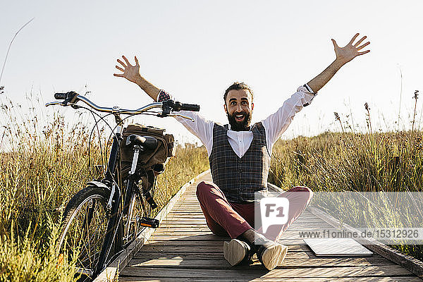Glücklicher  gut gekleideter Mann sitzt auf einem Holzsteg auf dem Land neben einem Fahrrad