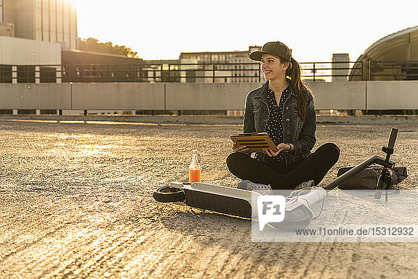 Lächelnde junge Frau mit Roller  Getränk und Tablette sitzt bei Sonnenuntergang auf dem Parkdeck