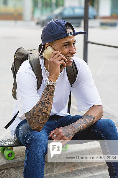 Portrait of tattooed young man on the phone sitting on his skateboard outdoors