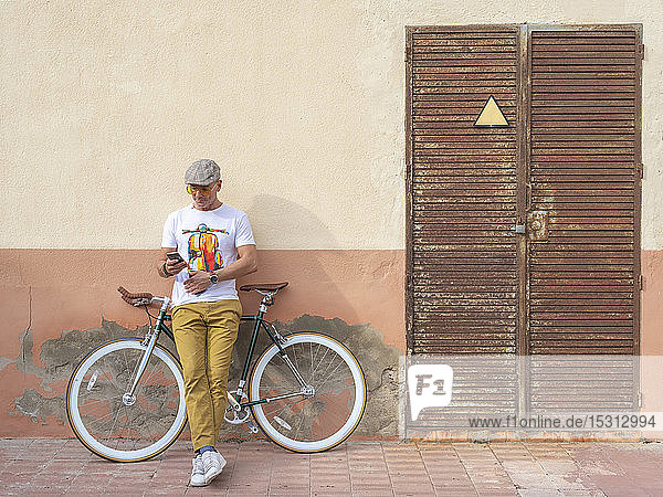 Mann mit Fixie-Fahrrad lehnt an Wand und sieht Handy an