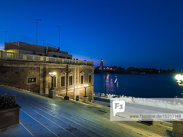 Illuminated Roman column on steps amidst buildings by sea in Brindisi against sky at night