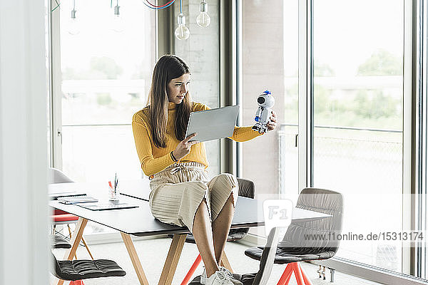Junge Geschäftsfrau sitzt im Büro auf dem Tisch und hält Roboter und Tablet