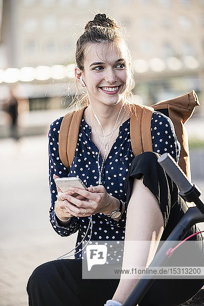 Lächelnde junge Frau mit Elektroroller  Kopfhörern und Mobiltelefon
