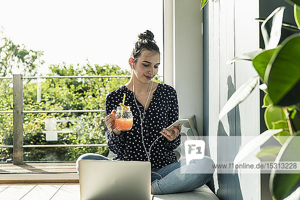 Lächelnde junge Frau mit Laptop  Handy und gesundem Getränk zu Hause