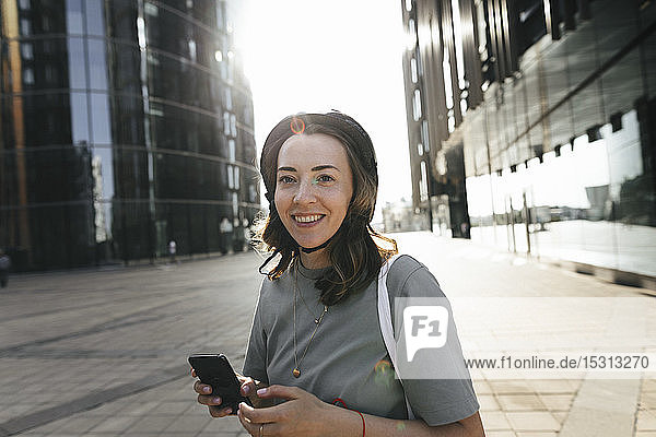 Frau mit einem Helm für einen E-Scooter mit Smartphone  im Hintergrund moderne Bürogebäude