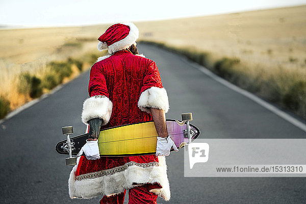 Rückansicht des Weihnachtsmanns mit einem Longboard in der Hand auf der Landstraße