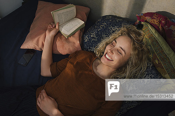 Portrait of laughing young woman with eyes closed lying on bed with a book