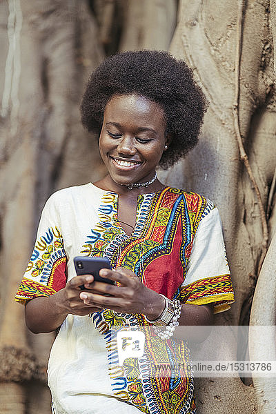 Lächelnde junge Frau  die sich mit ihrem Smartphone an einen Baumstamm lehnt