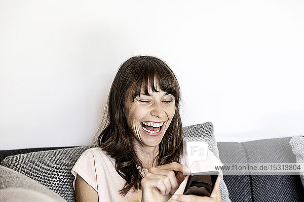 Lachende Frau benutzt Smartphone auf der Couch zu Hause