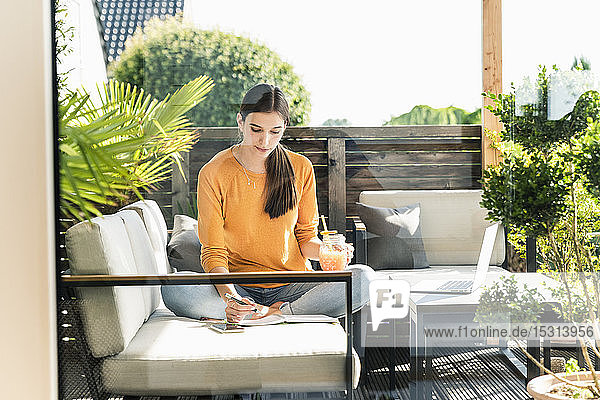 Junge Frau sitzt auf einer Couch auf der Terrasse mit einem gesunden Getränk und einem Notizbuch