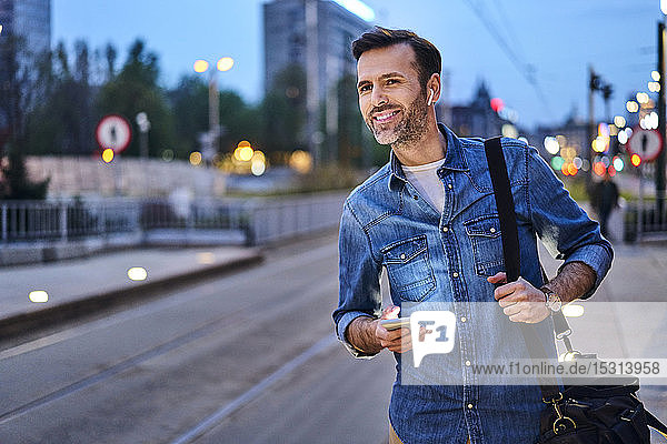 Lächelnder Mann mit drahtlosen Kopfhörern und Smartphone wartet an der Straßenbahnhaltestelle während des abendlichen Pendelns nach der Arbeit
