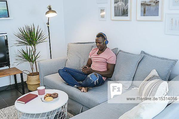 Junger Mann mit Kopfhörern und Smartphone auf der Couch zu Hause