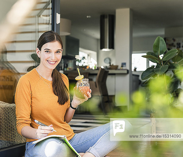 Porträt einer lächelnden jungen Frau mit gesundem Getränk und Notizbuch an der Terrassentür