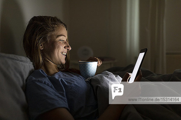 Young woman lying in bed at home with cup of tea at night using tablet
