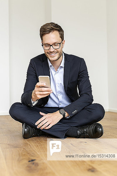 Lächelnder Geschäftsmann sitzt auf dem Boden und benutzt sein Handy