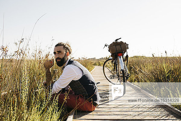 Gut gekleideter Mann  der auf einem Holzsteg auf dem Land neben einem Fahrrad sitzt