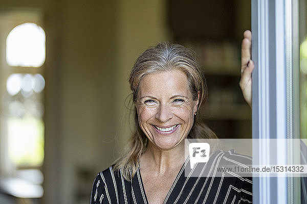 Porträt einer lächelnden reifen Frau  die an einer geöffneten Terrassentür steht