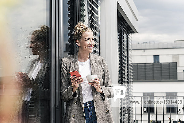 Lächelnde Geschäftsfrau mit Handy und Kaffeetasse auf der Dachterrasse stehend