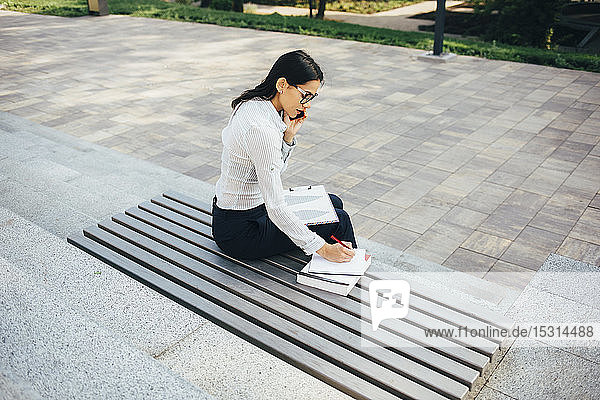 Young businesswoman sitting on a bench talking on cell phone and taking notes