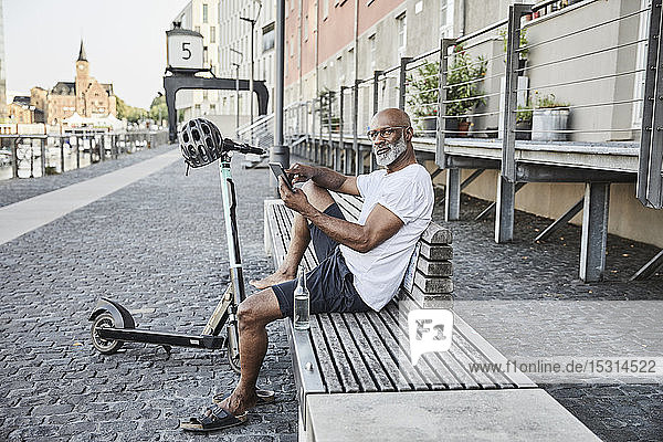 Portrait eines reifen Mannes mit einem E-Scooter  der sich im Sommer auf einer Bank entspannt und sein Handy benutzt  Köln  Deutschland