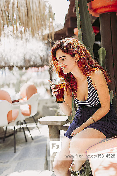 Glückliche junge Frau mit einem Getränk auf einer Bank im Freien sitzend