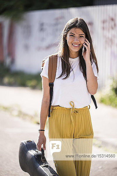 Portrait of smiling young woman carrying violin case talking on the phone on the street