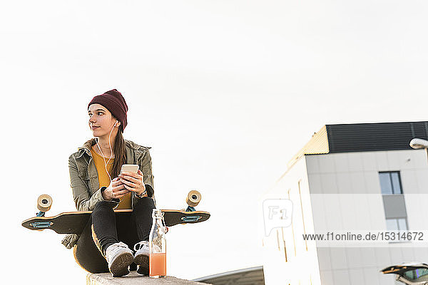 Stilvolle junge Frau mit Skateboard und Handy an der Wand sitzend