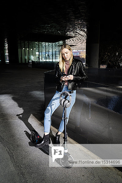 Young woman with kick scooter and headphones using smartphone