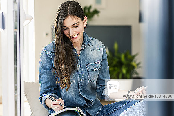 Smiling young woman wearing denim shirt taking notes at home