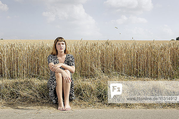 Portrait of young woman sitting barefoot at roadside in front of grain field