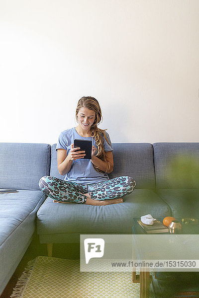 Smiling young woman sitting on couch at home using tablet