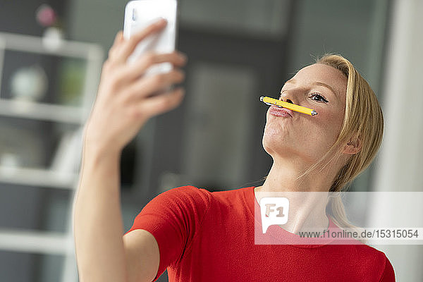 Verspielte junge Frau balanciert im Büro Stift auf dem Mund und nimmt ein Selfie