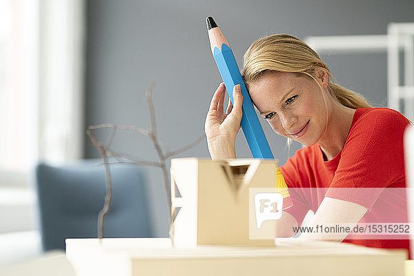 Junge Frau im Büro mit übergroßem Stift und Architekturmodell auf dem Schreibtisch