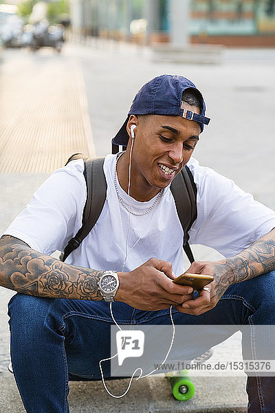 Portrait of tattooed young man sitting on skateboard listening music with smartphone and earphones