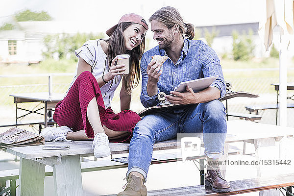 Junges Paar sitzt am Tisch in einem Biergarten mit Tablette  Sandwich und Kaffee zum Mitnehmen
