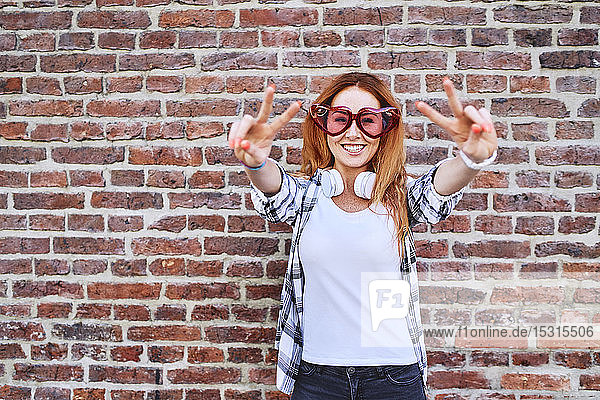 Fröhliche junge Frau mit herzförmiger Brille macht Friedensgesten und lächelt in die Kamera