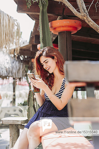 Junge Frau mit einem Getränk und Handy auf einer Bank im Freien sitzend